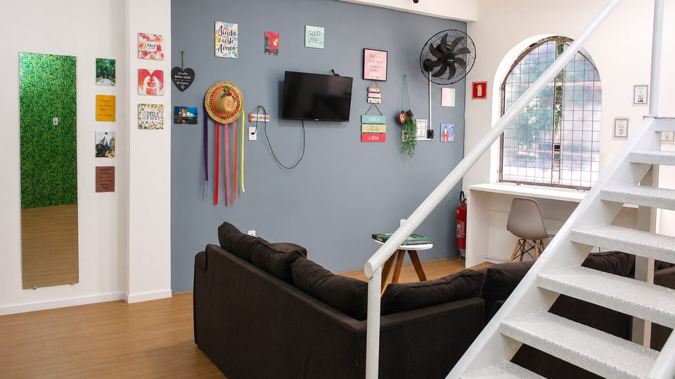 Área comum do hostel, com sofá, escada branca e parede de jardim vertical.