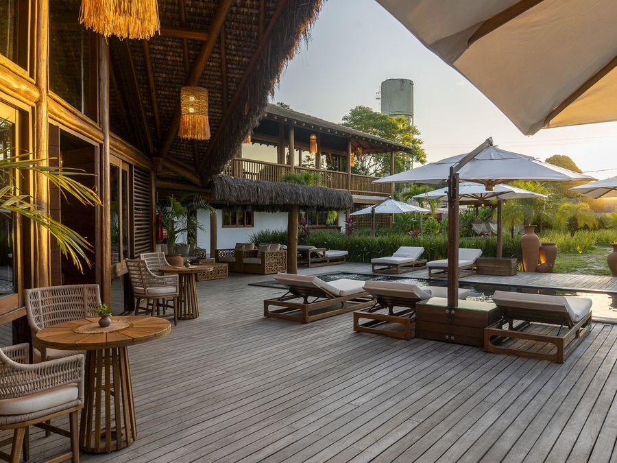 Deck de madeira com mesas, espreguiçadeiras e a piscina ao fundo no fim da tarde.
