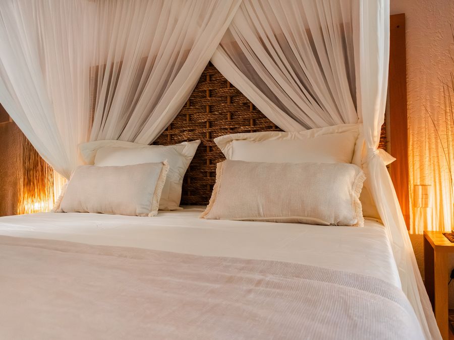 Cama de casal com mosquiteiro branco e abajur aceso ao lado.